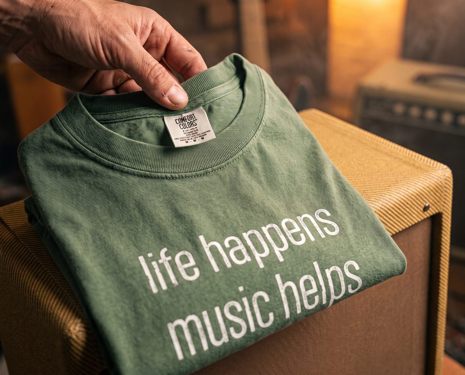 Hand opening the collar of the moss green Life Happens Music Helps tee to show the Comfort Colors neck label, on a tweed amp