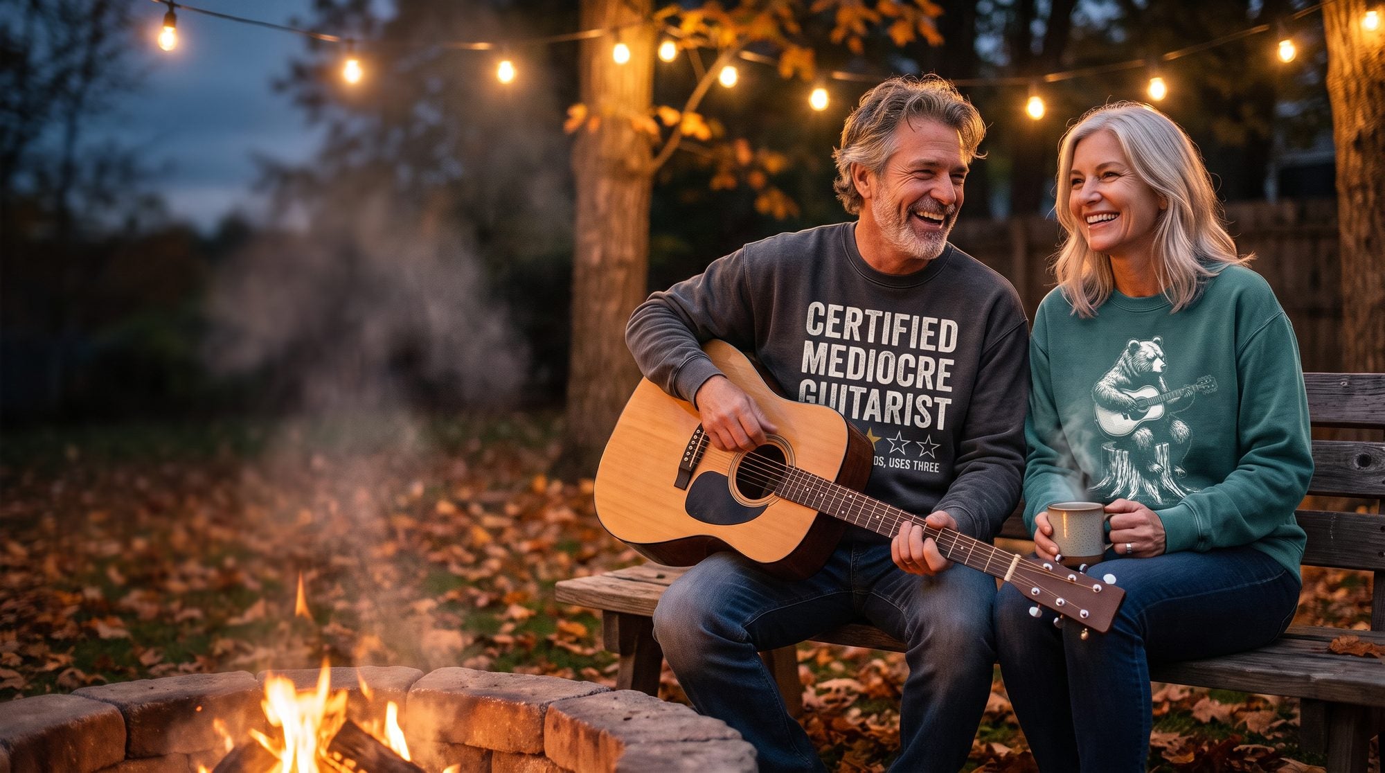 Couple laughing by a backyard fire pit in Fret Folk sweatshirts: Certified Mediocre Guitarist and Bear Guitarist, he plays an acoustic guitar